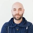 Headshot of a bald man with a dark beard wearing a blue hooded sweater.